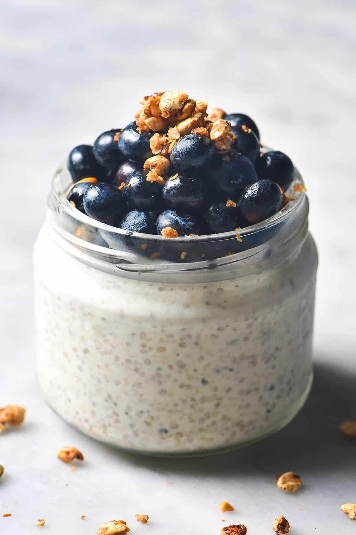 A close up side on image of a round jar filled with gluten free overnight oats and topped with blueberries and a sprinkle of granola. The jar sits on a white marble table and a sprinkle of granola surrounds the jar. 