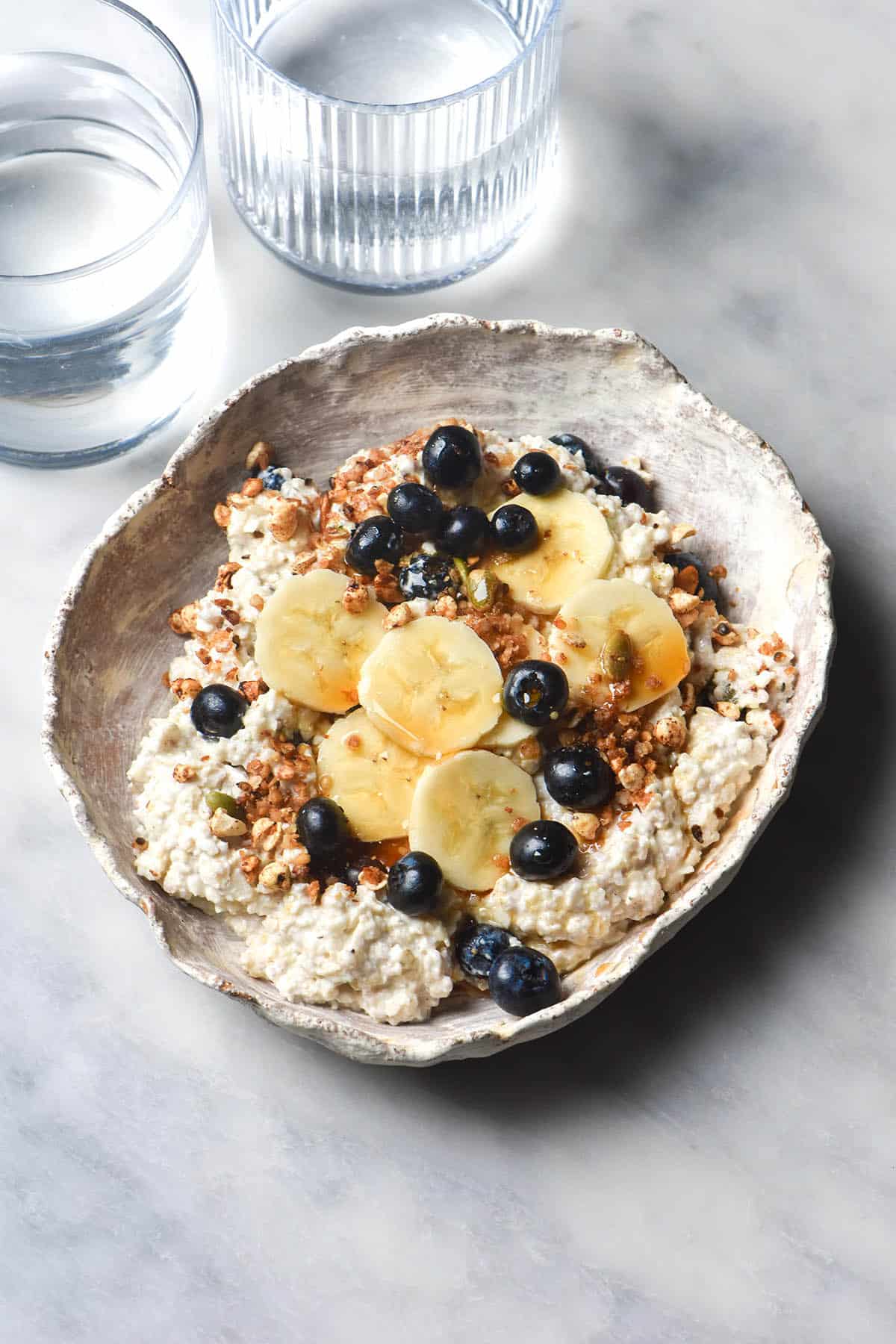 An aerial image of gluten free overnight oats that have been decanted in a textured white ceramic bowl and topped with banana slices, blueberries and maple syrup. The bowl sits atop a white marble table and two sunlit water glasses sit to the top left of the image.