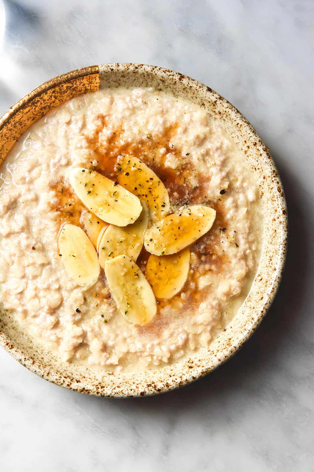 An aerial image of a speckled beige ceramic bowl filled with gluten free porridge that has been topped with banana coins, maple syrup and hemp seeds. The porridge sits atop a white marble table with water glasses off to the left hand side of the image