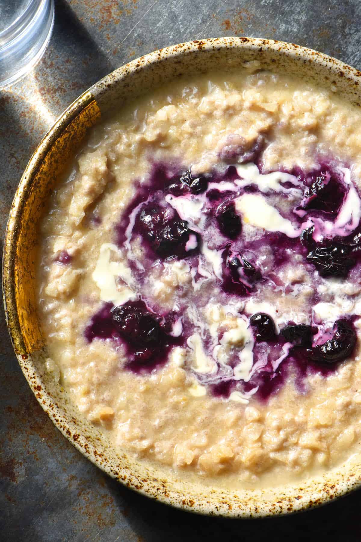A close up aerial view of a bowl of gluten free porridge topped with blueberry compote and swirled with cream. The wide mouthed ceramic bowl sits atop a dark blue steel backdrop with a glass of water in the top left corner