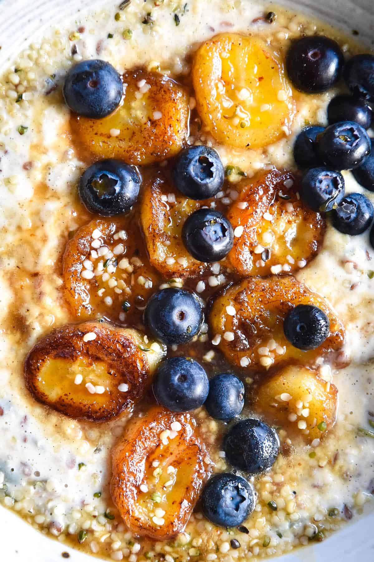 An aerial close up view of a bowl of seedy gluten free porridge topped with caramelised bananas, fresh blueberries and a sprinkle of hemp seeds