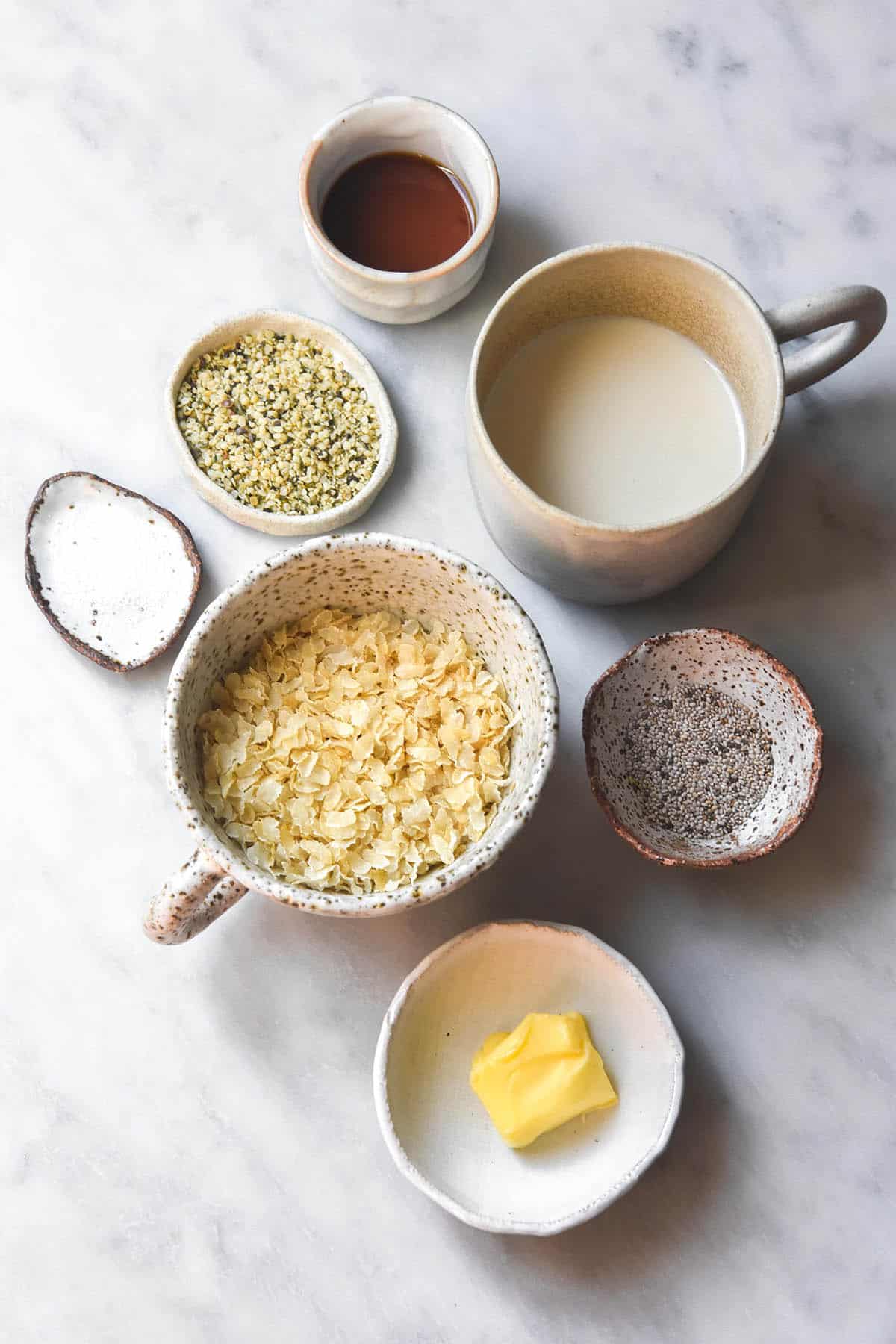 An aerial image of the ingredients used for gluten free porridge in small white ceramic bowls arranged casually on a white marble table. 
