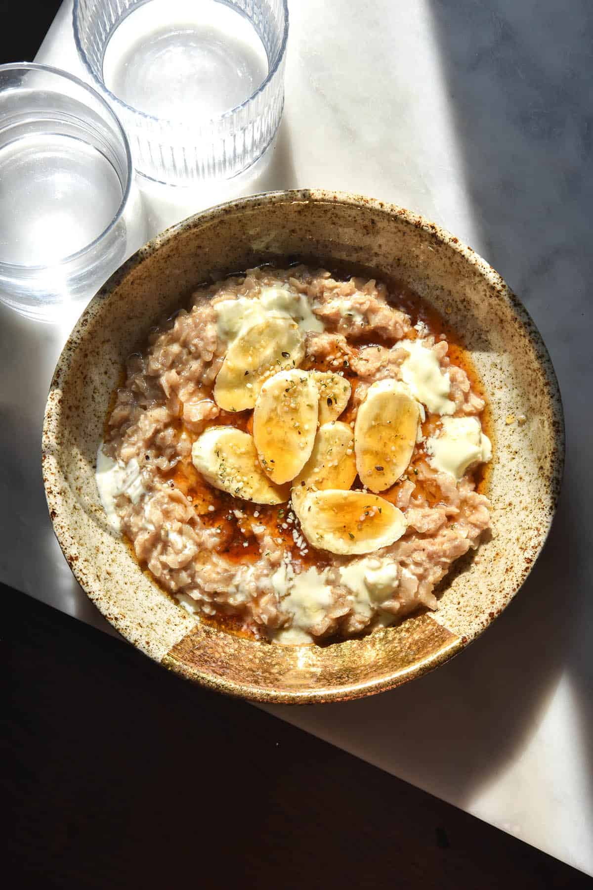 An aerial sunlit image of a ceramic bowl filled with gluten free porridge and topped with banana coins, hemp seeds, maple syrup and yoghurt. The bowl sits in bright contrasting light and two glasses of water sit to the top left of the image. 