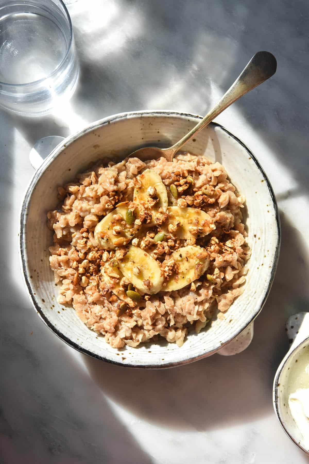 An aerial sunlit image of a bowl of gluten free porridge topped with banana coins and gluten free granola. The bowl sits atop a white marble table and two glasses of water sit to the top left of the image
