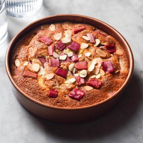 An aerial side on image of a gluten free rhubarb cake in a pale pink cake tin atop a white marble table. The cake is golden brown and topped with pink rhubarb and almond flakes.