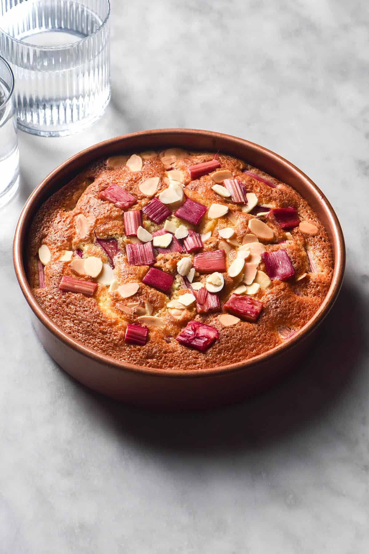 An aerial side on image of a gluten free rhubarb cake in a pale pink cake tin atop a white marble table. The cake is golden brown and topped with pink rhubarb and almond flakes.