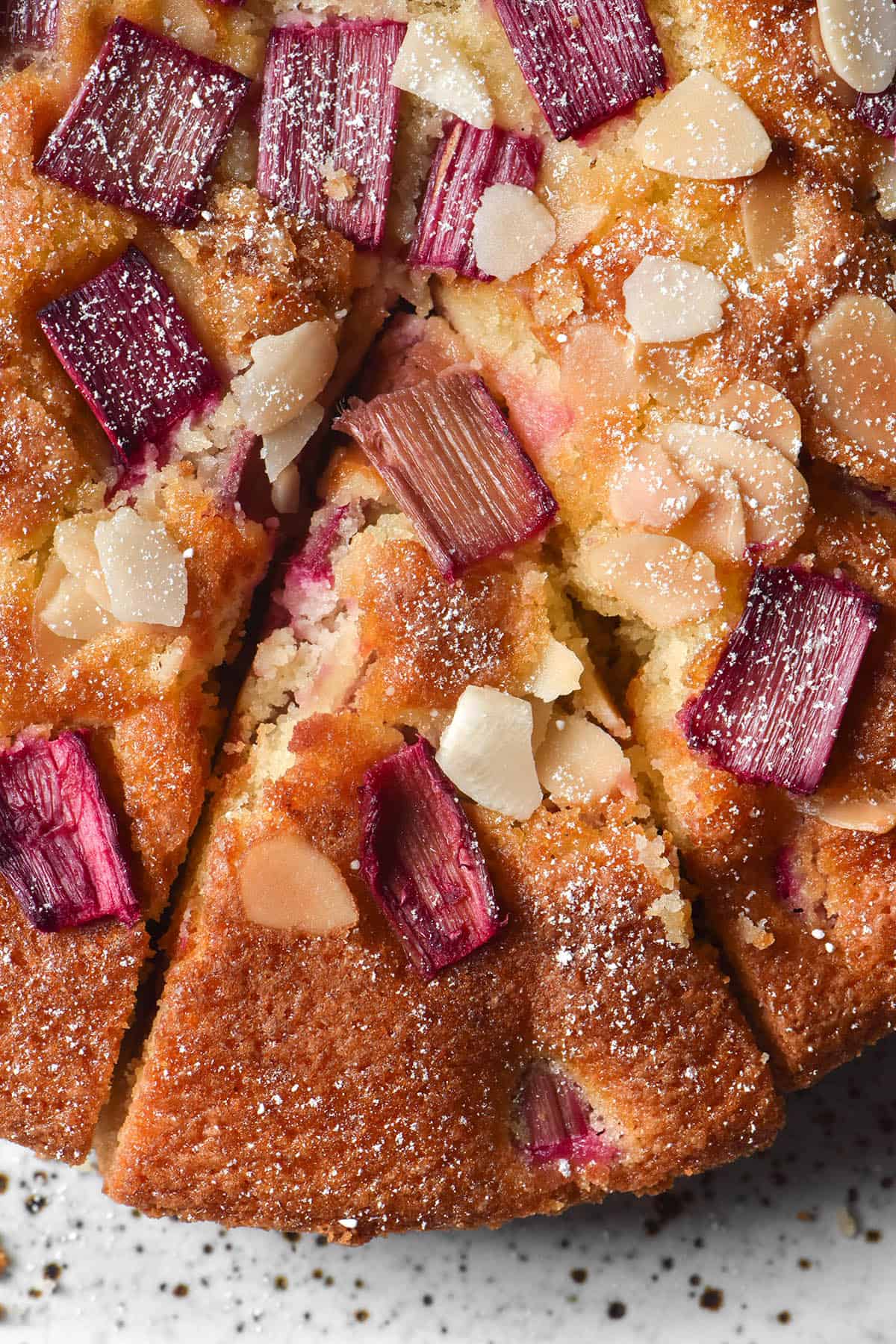 An aerial image of a gluten free rhubarb cake topped with icing sugar and flaked almonds. The cake is golden brown and sits atop a white speckled ceramic plate. The centre of the image is a slice about to be taken from the cake