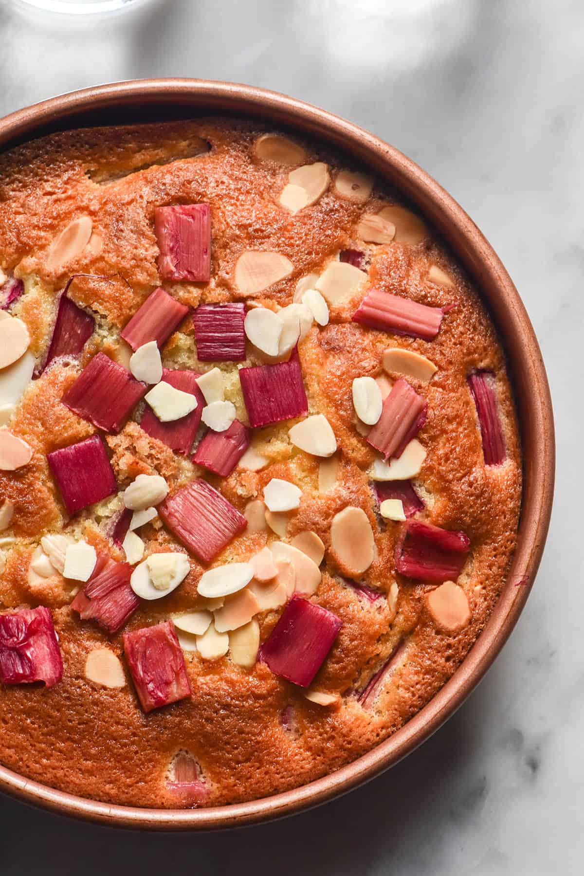 An aerial image of a gluten free rhubarb cake in a light pink cake tin atop a white marble table. The cake is golden brown and flecked with pink rhubarb pieces and flaked almonds.