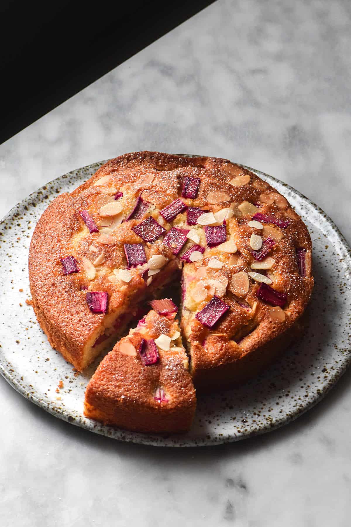 An aerial, side on image of a gluten free rhubarb cake on a white speckled plate atop a white marble table. The cake is golden brown and flecked with pink rhubarb and golden flaked almonds. It has been finished with a sprinkling of icing sugar. 