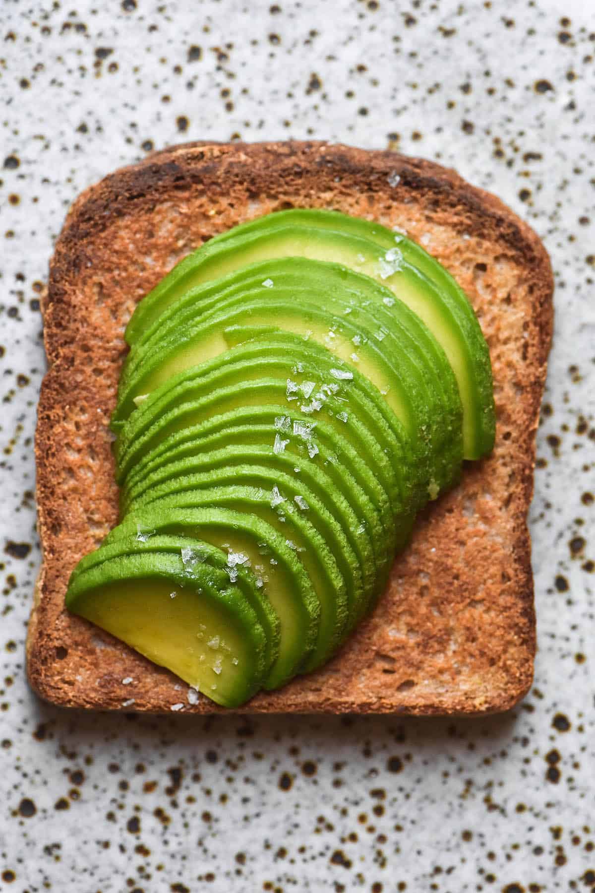 An aerial close up image of thinly sliced avocado atop a slice of gluten free sandwich bread. The bread sits on a white speckled ceramic plate and the avocado is topped with a thin line of flaky sea salt.