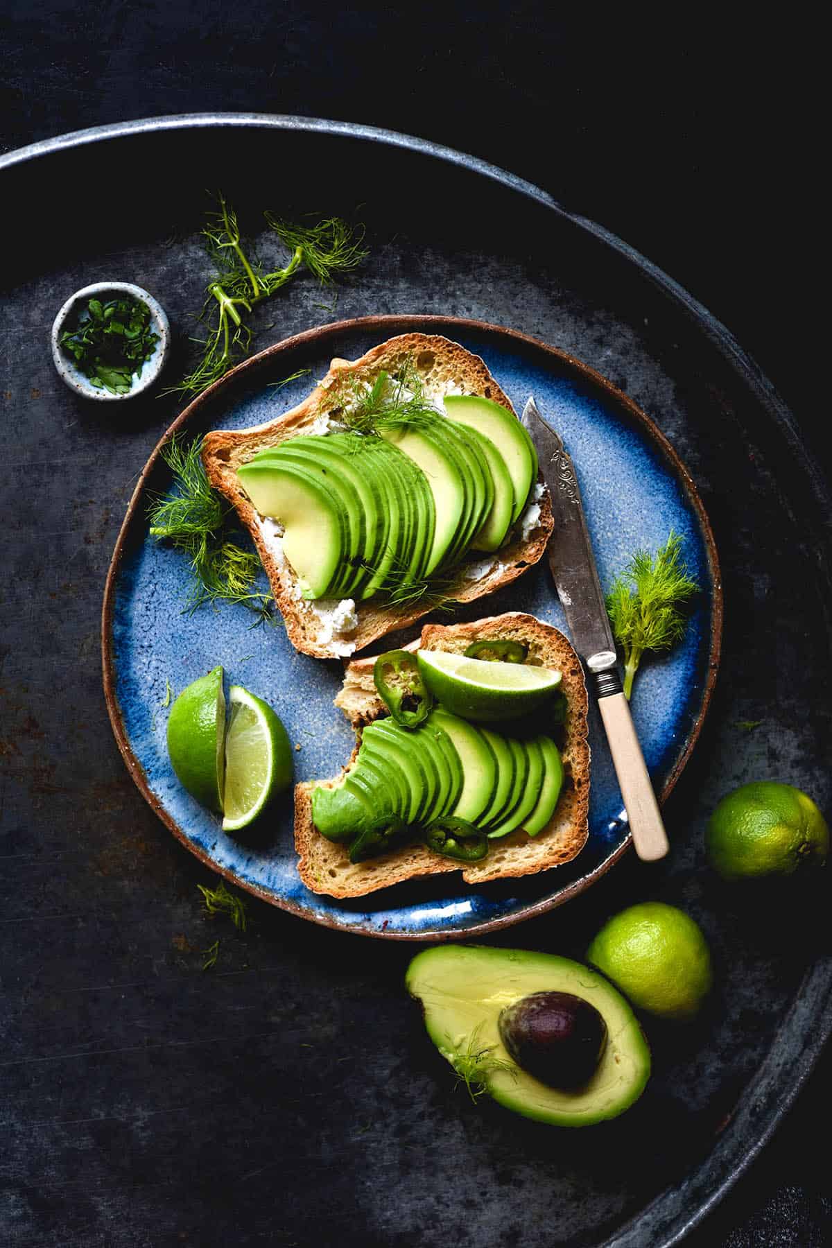 A moody aerial image of gluten free sandwich bread toasted and topped with goat's cheese and sliced avocado. The toast sits on a bright blue plate atop a moody dark steel backdrop, surrounded by extra avocados, herbs and limes