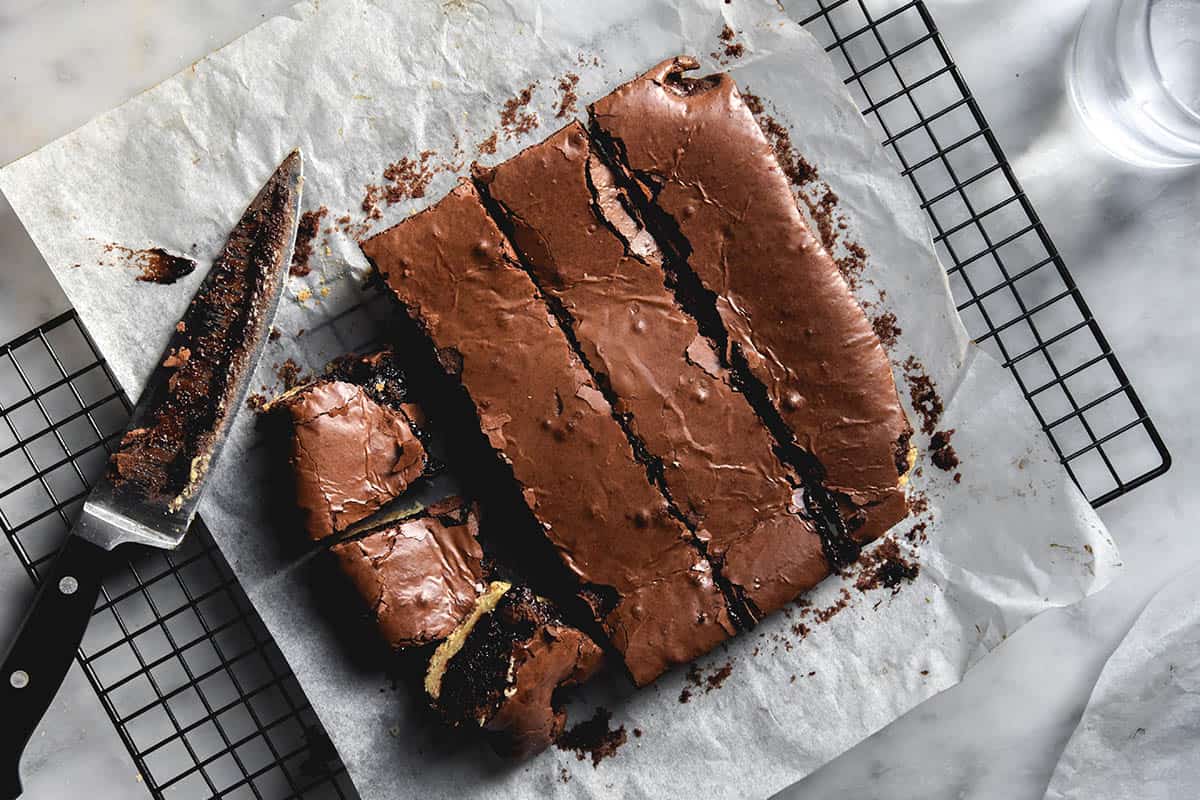 An aerial view of a slab of gluten free shortbread brownies on a sheet of baking paper atop a cooling rack on a white marble table