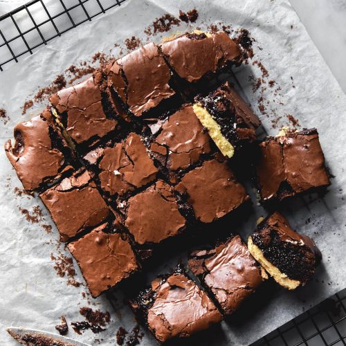 An aerial view of gluten free shortbread brownies on a sheet of baking paper on a wire cooling rack atop a white marble table. Two glasses of water sit to the top left of the image