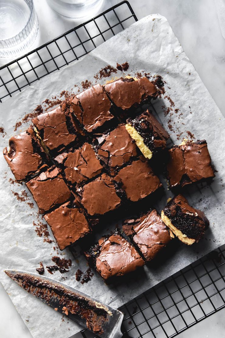 An aerial view of gluten free shortbread brownies on a sheet of baking paper on a wire cooling rack atop a white marble table. Two glasses of water sit to the top left of the image