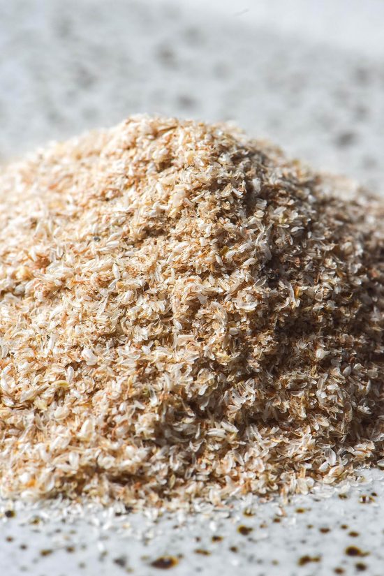 A close up macro image of a pile of psyllium husk flakes on a white speckled ceramic plate