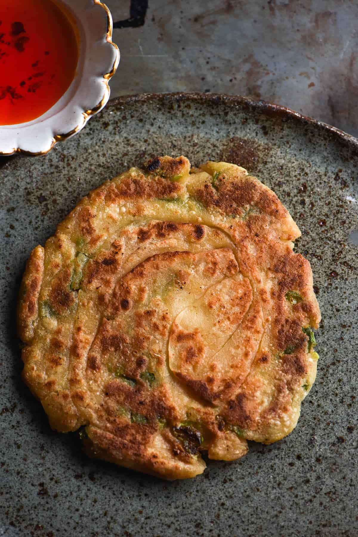 An aerial image of a gluten free, low FODMAP scallion pancake on a speckled dark blue ceramic plate. A small bowl of low FODMAP chilli oil sits in the top lefthand corner.