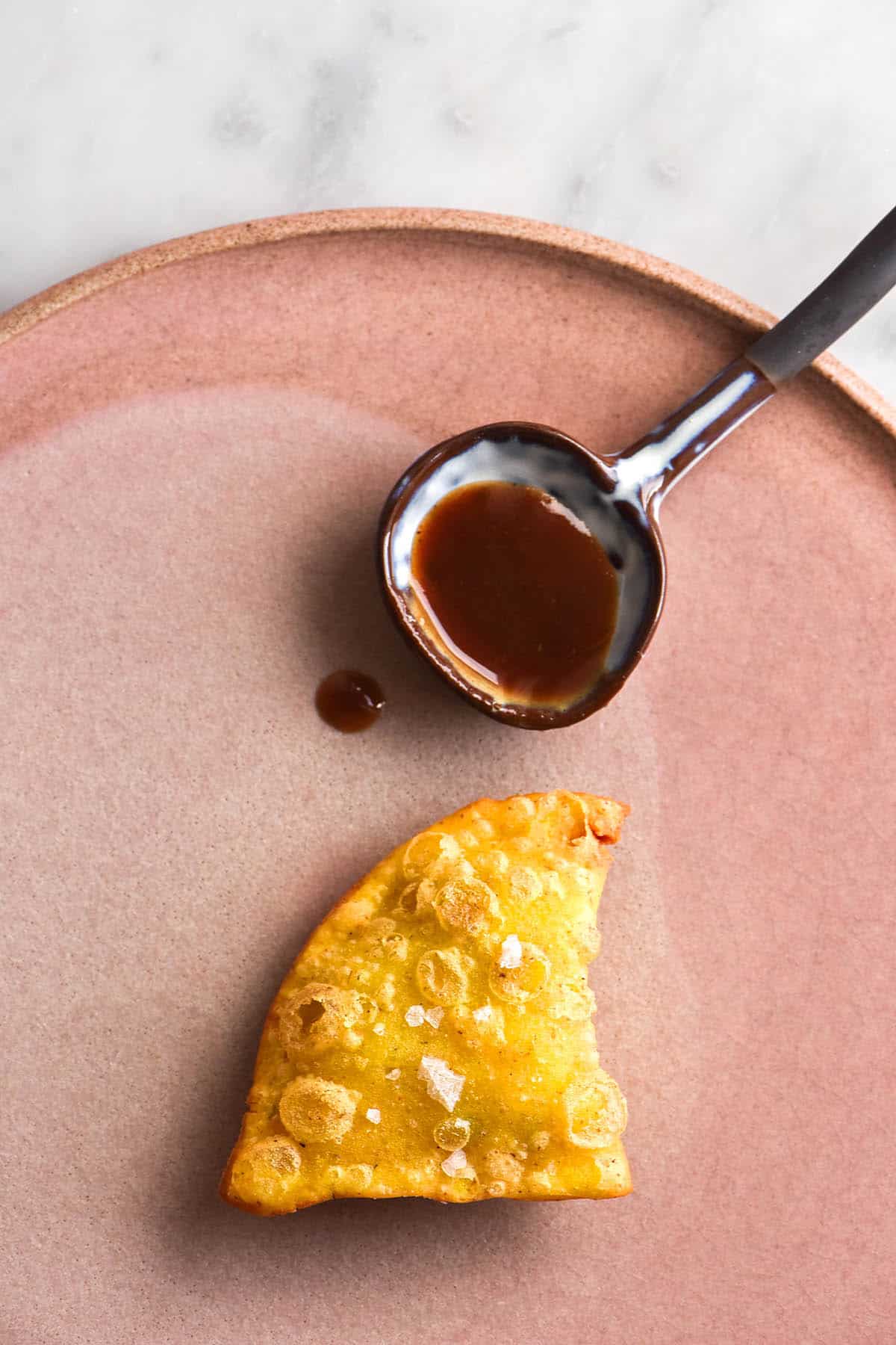 An aerial image of a crispy fried gluten free samosa on a pale pink ceramic plate. Above the samosa sits a ceramic spoon with imli (tamarind) chutney