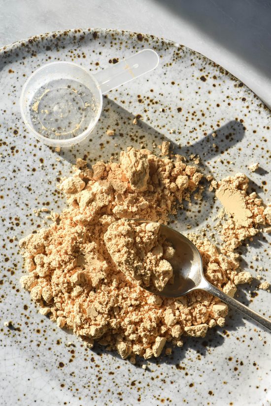 An aerial sunlit view of a scoop of peanut protein on a white speckled ceramic plate. A spoon digs into the protein and the scoop sits empty above the powder.