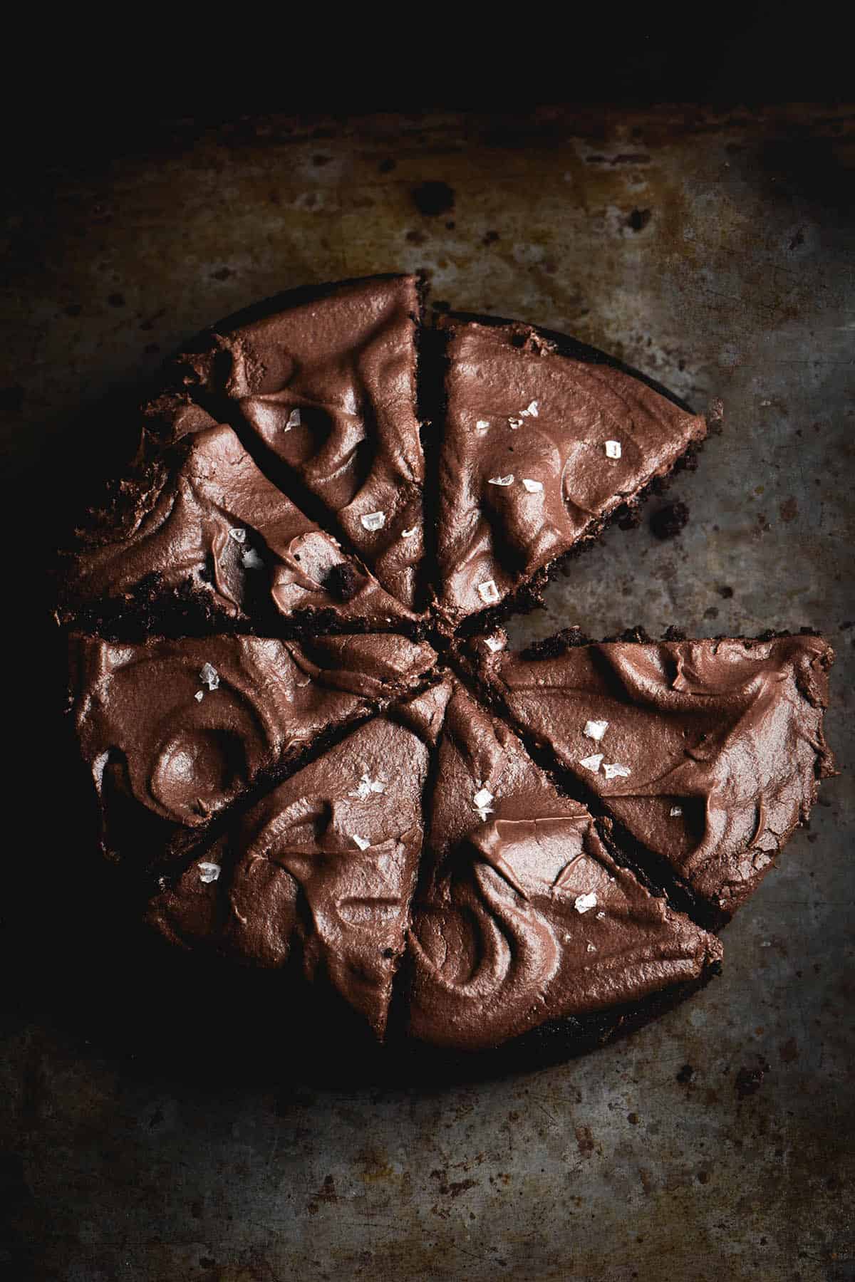 An aerial image of a gluten free vegan chocolate cake topped with chocolate buttercream icing. The cake has been sliced and the buttercreamed topped with some sea salt flakes. It sits atop a dark metal mottled backdrop.
