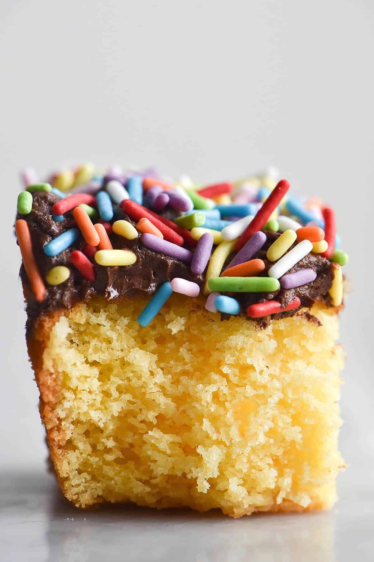 A side on macro image of a slice of vanilla birthday cake on a white marble table against a white backdrop. The cake is topped with chocolate buttercream and rainbow sprinkles. 