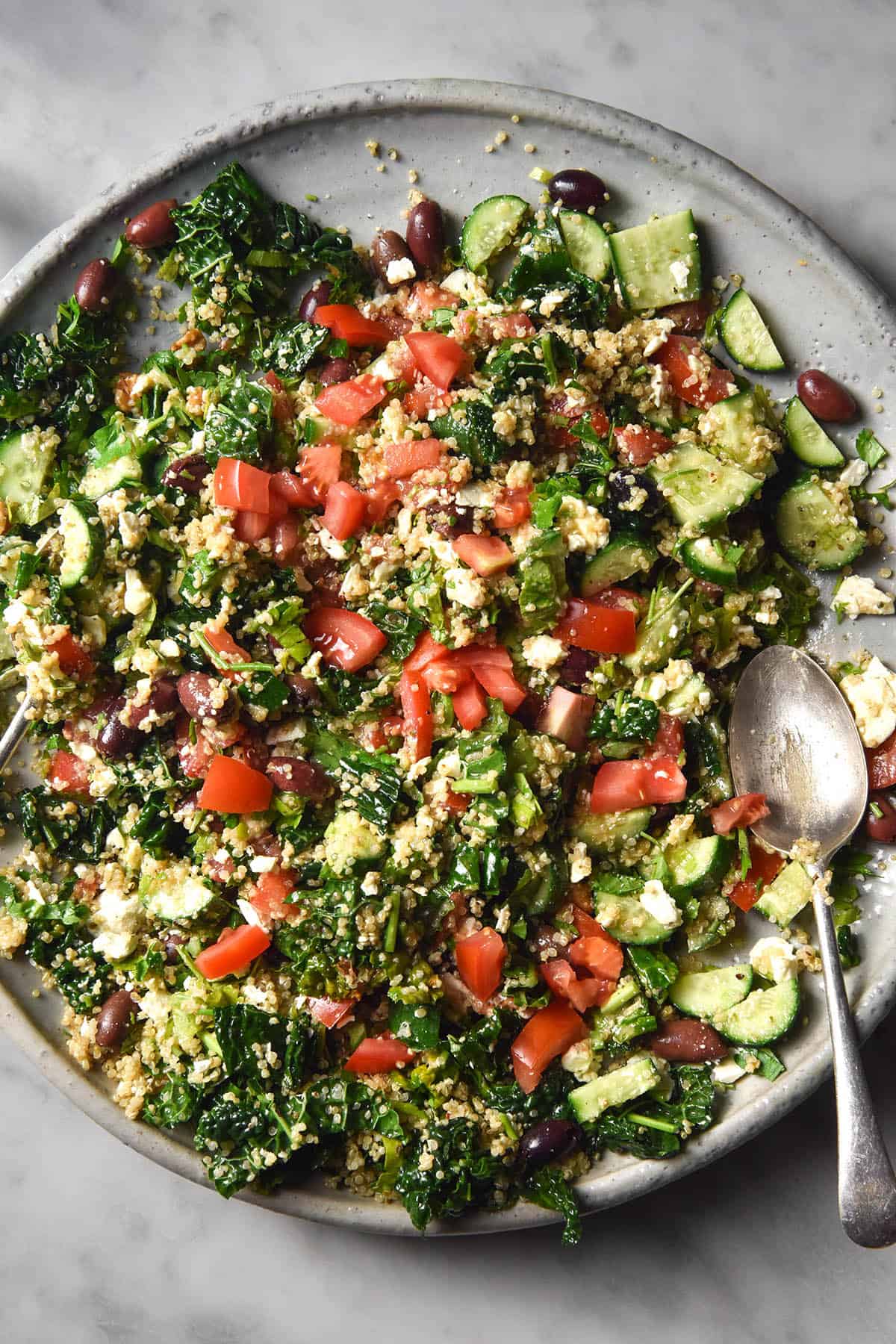 An aerial close up image of a quinoa, tomato, cucumber herb and feta salad with olives. The salad sits atop a large ceramic platter on a white marble table