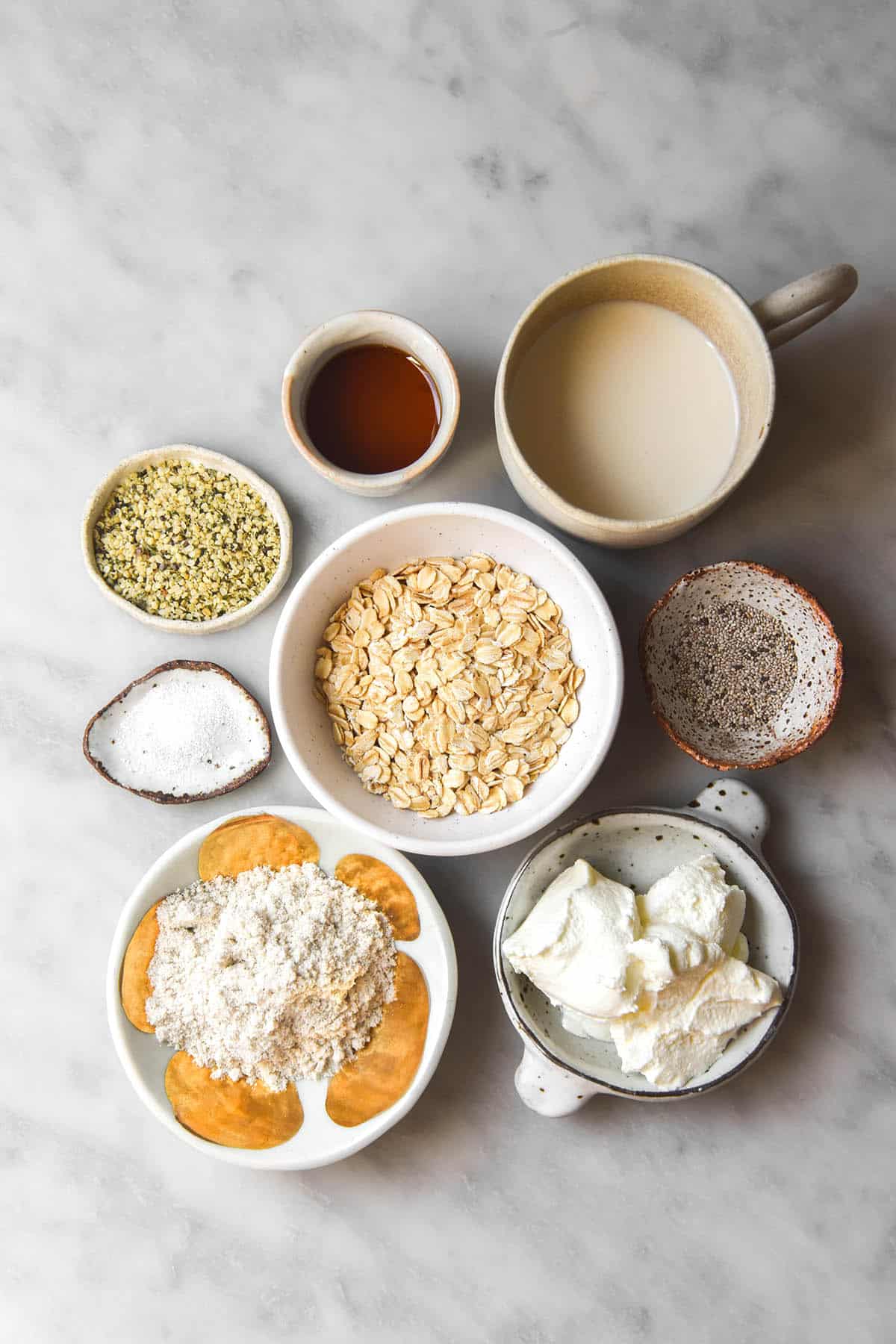 An aerial image of the ingredients used to make low FODMAP overnight oats. The ingredients are all in small white ceramic bowls arranged casually on a white marble table. 