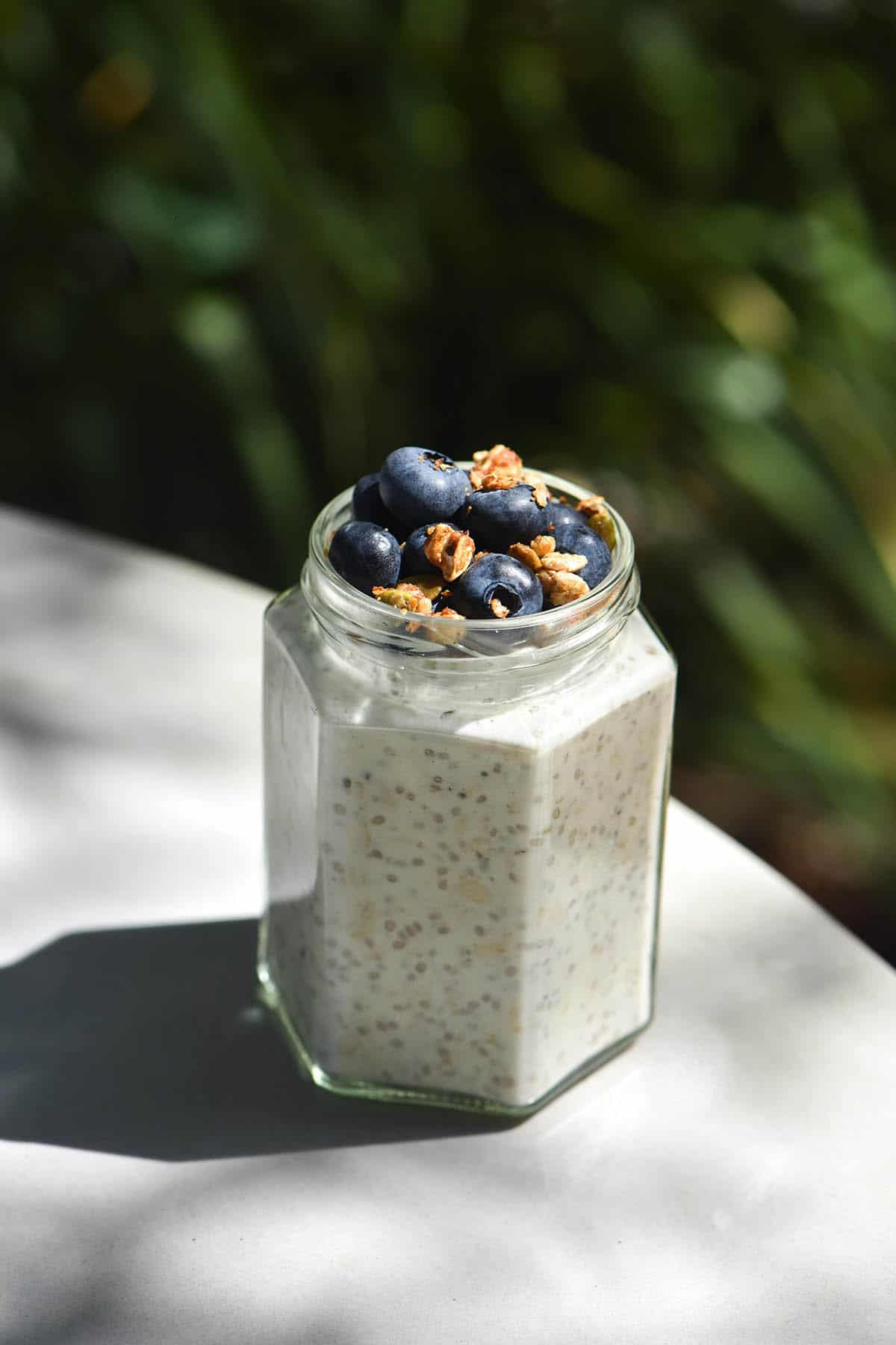 A side on image of a jar of low FODMAP overnight oats in bright dappled sunlight. The jar is topped with blueberries and granola and sits on a grey outdoor table in front of a leafy green bokeh background