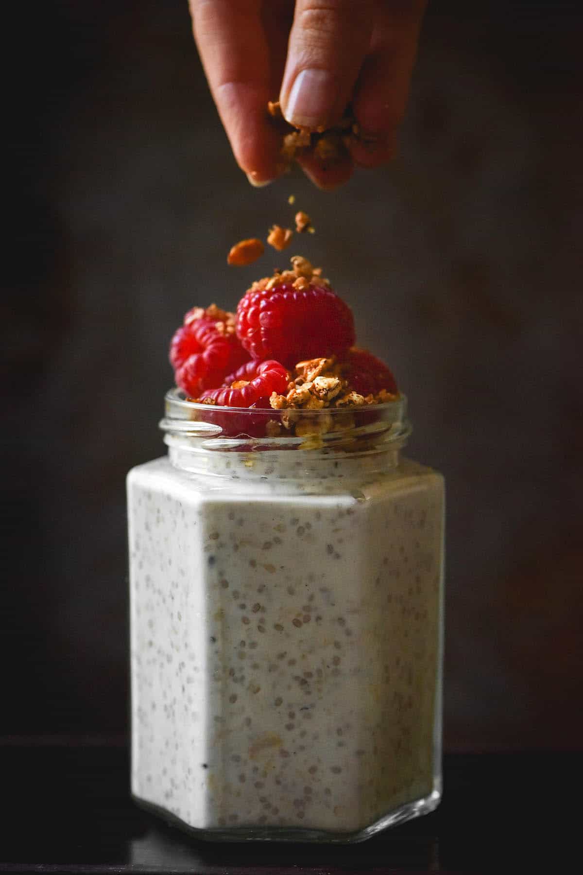 A side on moody image of a jar of low FODMAP overnight oats topped with raspberries and granola. A hand extends down from the top of the image to sprinkle extra granola atop the raspberries 