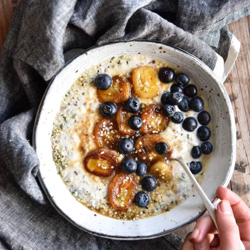 An aerial image of a bowl of low FODMAP overnight oats topped with caramelised bananas and fresh blueberries. The bowl sits on a wooden table and a denim blue linen tablecloth. A hand sticks a spoon into the bowl from the bottom left hand side