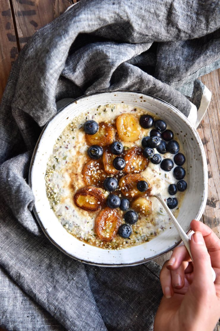 An aerial image of a bowl of low FODMAP overnight oats topped with caramelised bananas and fresh blueberries. The bowl sits on a wooden table and a denim blue linen tablecloth. A hand sticks a spoon into the bowl from the bottom left hand side