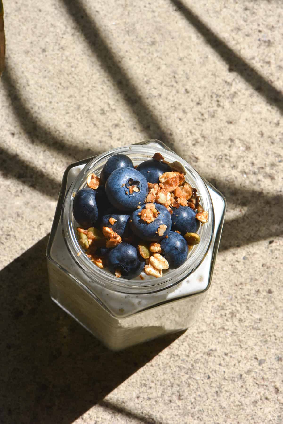 An aerial image of a jar of low FODMAP overnight oats topped with blueberries and extra granola. The jar sits on a stone paver and the contrasting sunlight and shadow cast by a plant in the background creates a unique shadow pattern across the image