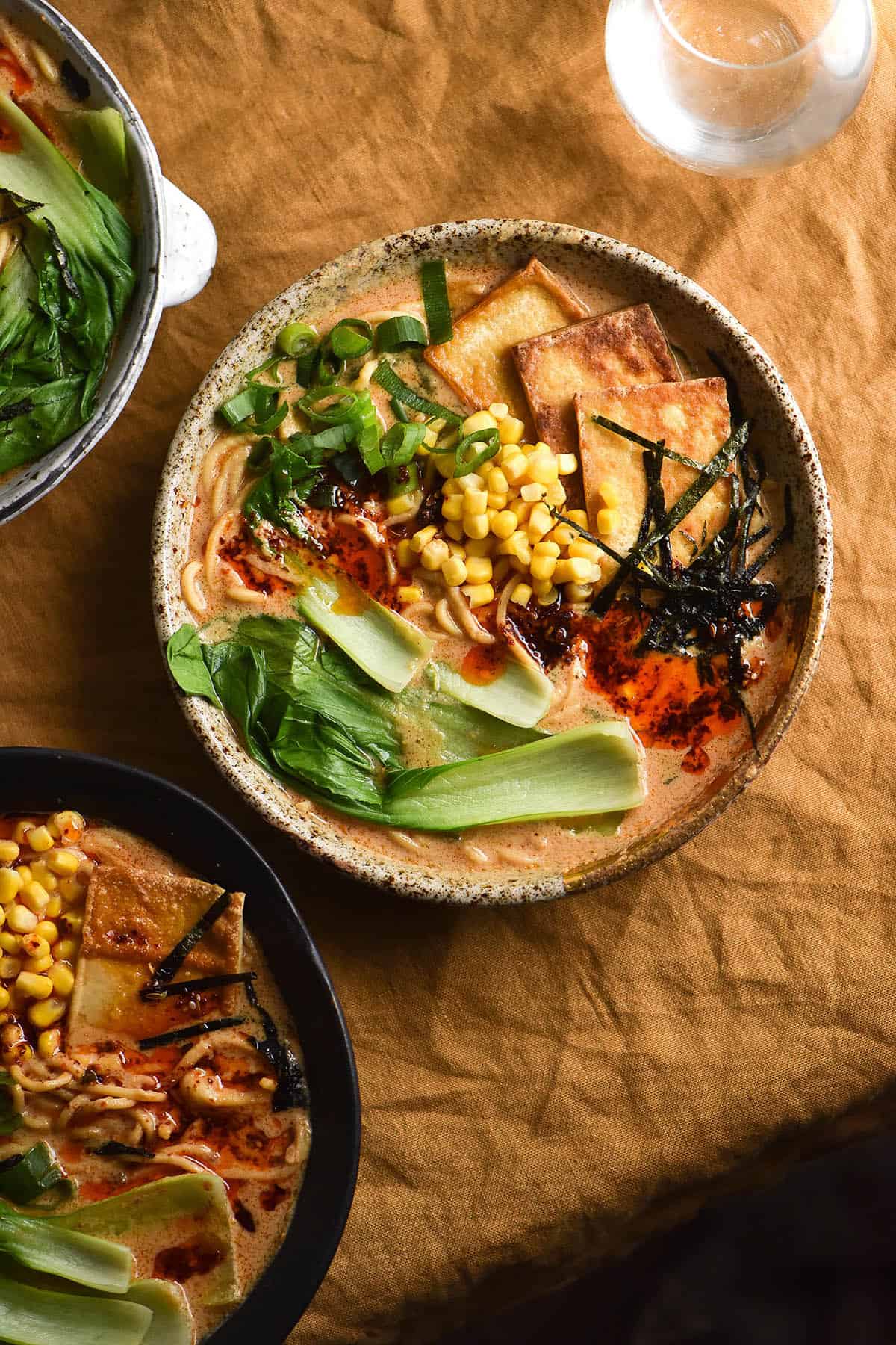 An aerial image of three bowls of ramen on a turmeric coloured tablecloth. The bowls are all different sizes and colours. The ramen is topped with tofu, sliced nori, chilli oil, bok choi, spring onion greens and corn kernels. 