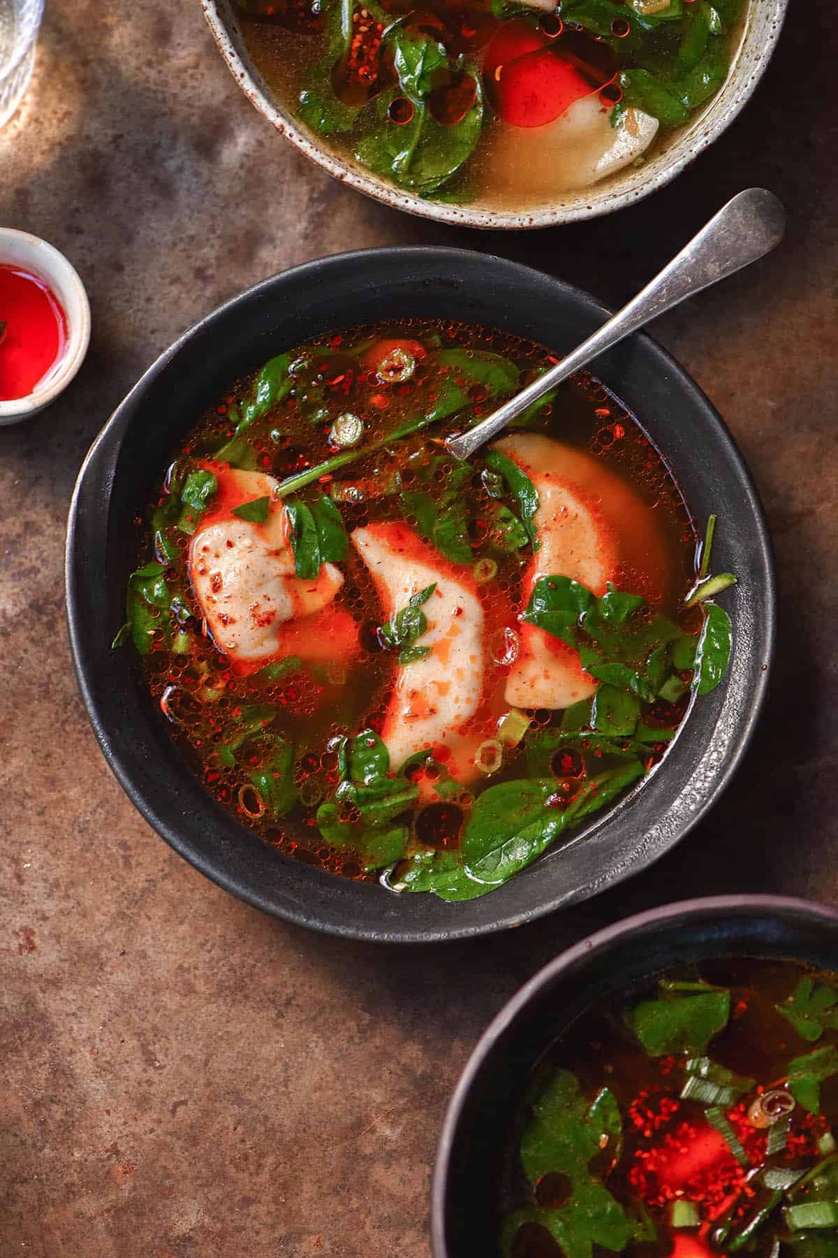 An aerial moody image of low FODMAP dumpling soup bowls on a rusty backdrop. The soup is topped with chilli oil and greens