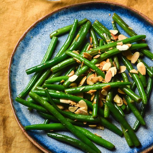 An aerial view of a blue ceramic plate topped with low FODMAP green beans and toasted almonds. The plate sits atop a mustard linen tablecloth and two water glasses sit to the top left of the image