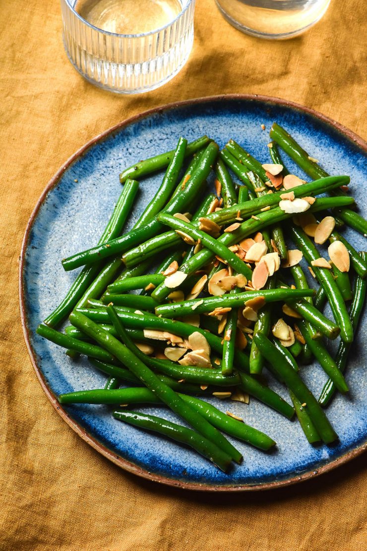 An aerial view of a blue ceramic plate topped with low FODMAP green beans and toasted almonds. The plate sits atop a mustard linen tablecloth and two water glasses sit to the top left of the image
