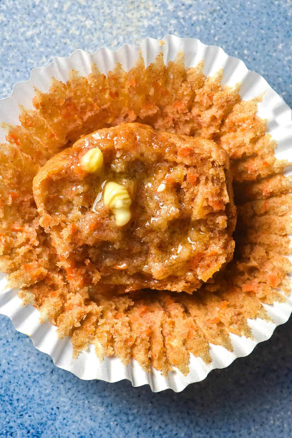 An aerial image of half a gluten free banana carrot muffin on a bright blue ceramic plate. The muffin half sits facing upwards on the muffin liner. It has been topped with melty butter