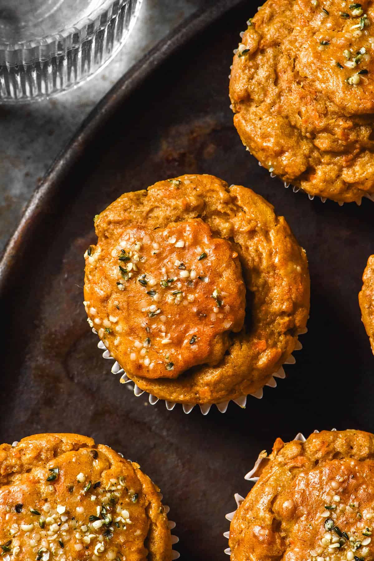 An aerial image of gluten free banana carrot muffins on a dark steel plate against a dark steel backdrop. A glass of water sits to the top left of the image