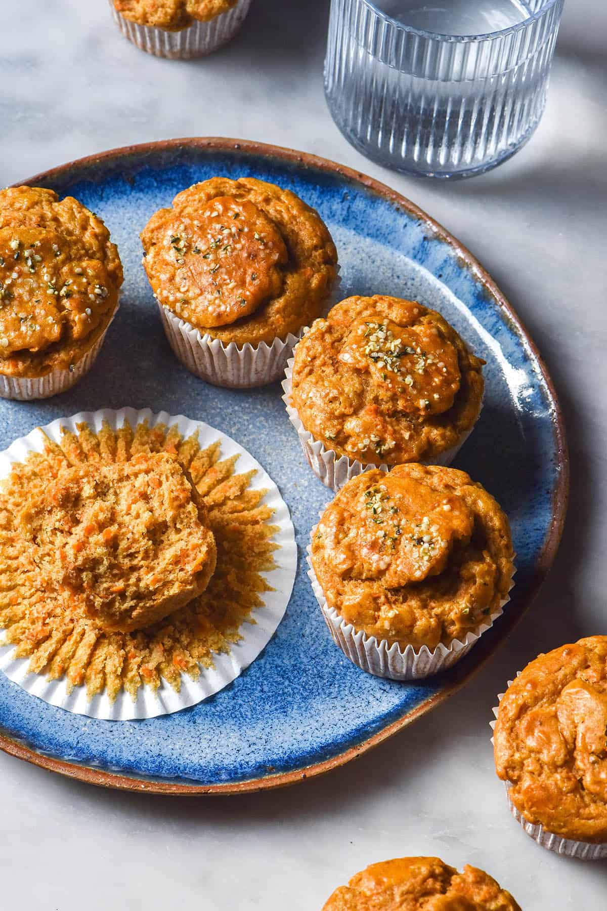 An aerial view of gluten free banana carrot muffins on a bright blue ceramic plate atop a white marble table. The muffins are casually arranged and a water glass sits to the top right of the image. 