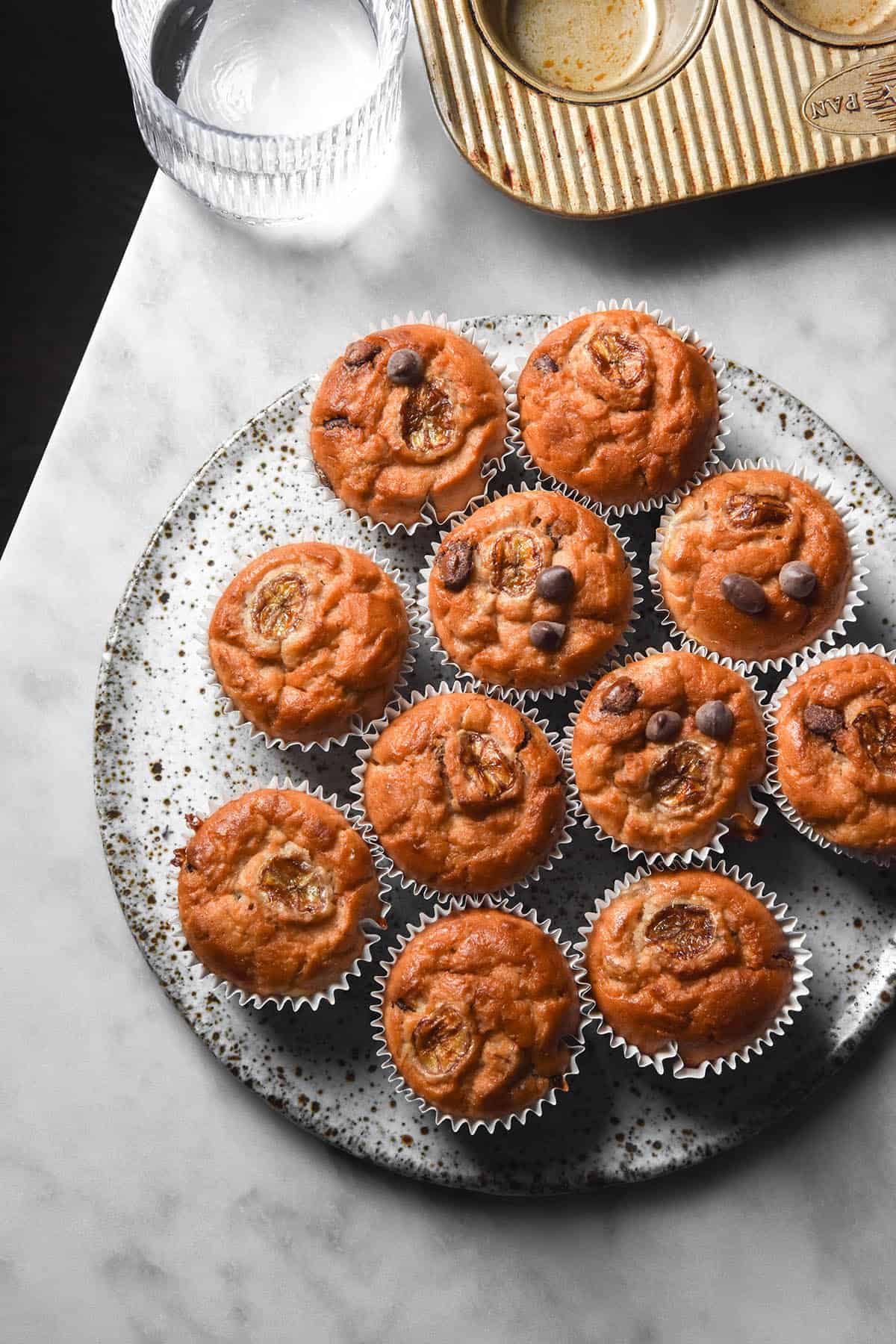 An aerial image of a white speckled ceramic plate topped with gluten free banana muffins. The plate sits on a white marble table and a glass of water and a muffin tray sit to the top of the image. 