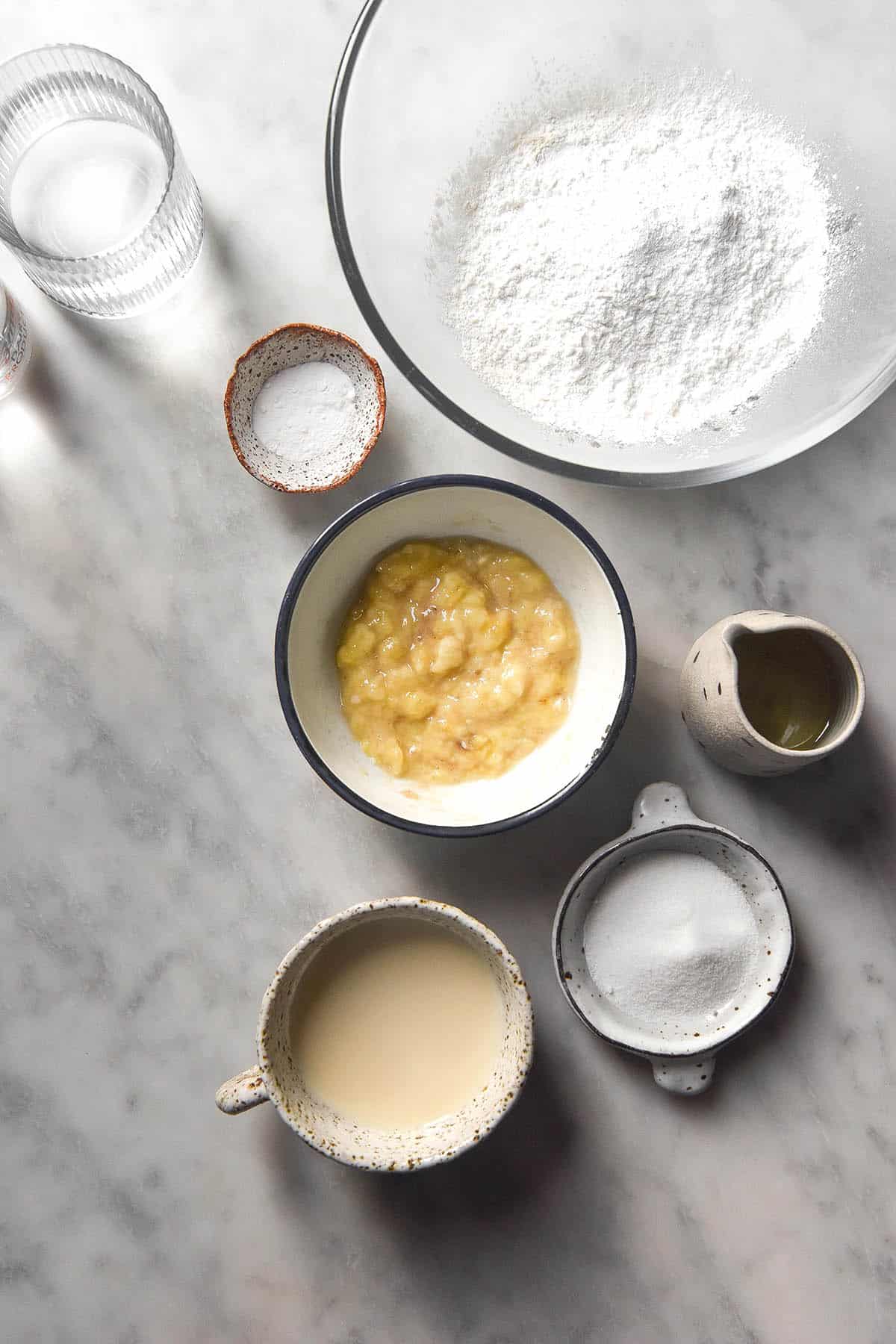 An aerial image of the ingredients used to make gluten free banana pancakes. The ingredients sit in various sized bowls on a white marble table.