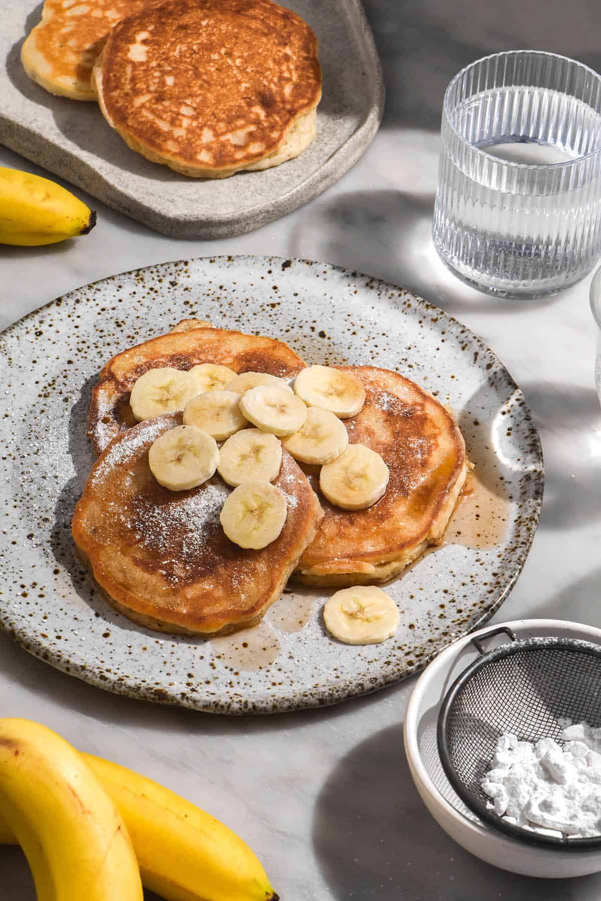 A side on view of a plate of white speckled ceramic plate topped with gluten free banana pancakes finished with banana slices and maple syrup. Water glasses, bananas and extra plates of pancakes surround the central plate. 