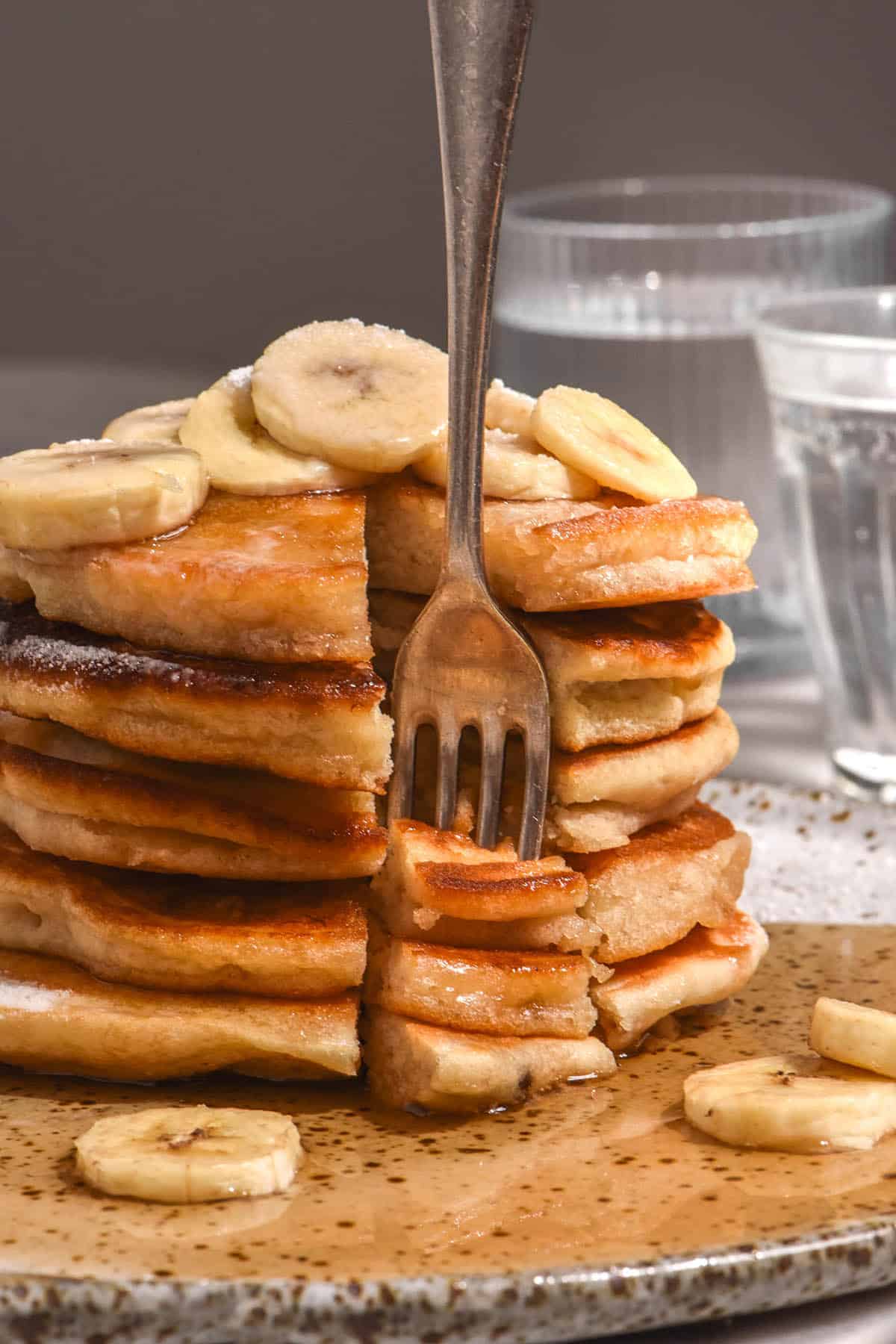 A side on view of a stack of gluten free banana pancakes that have been topped with banana slices and lots of maple syrup. A slice has been made into the stack in the front and a front stands upright in the pieces.