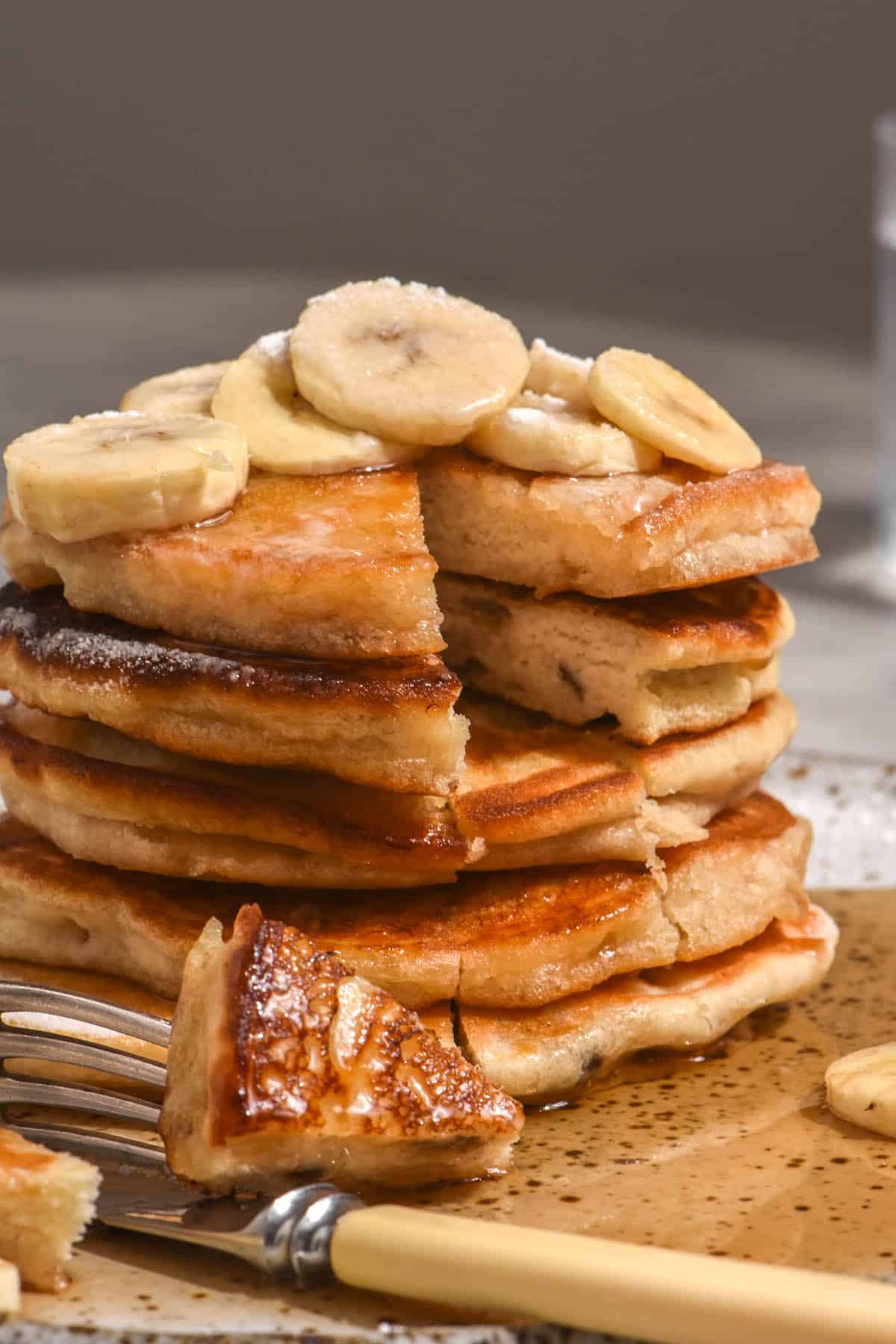 A side on view of a stack of gluten free banana pancakes that have been topped with banana slices and lots of maple syrup. A slice has been made into the stack in the front.
