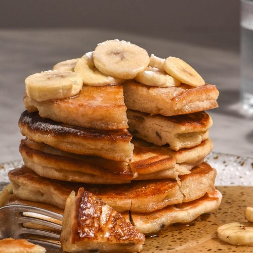 A side on view of a stack of gluten free banana pancakes that have been topped with banana slices and lots of maple syrup. A slice has been made into the stack in the front.