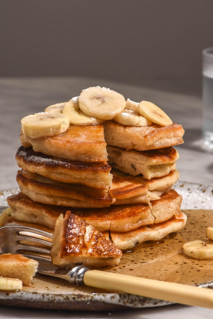 A side on view of a stack of gluten free banana pancakes that have been topped with banana slices and lots of maple syrup. A slice has been made into the stack in the front.