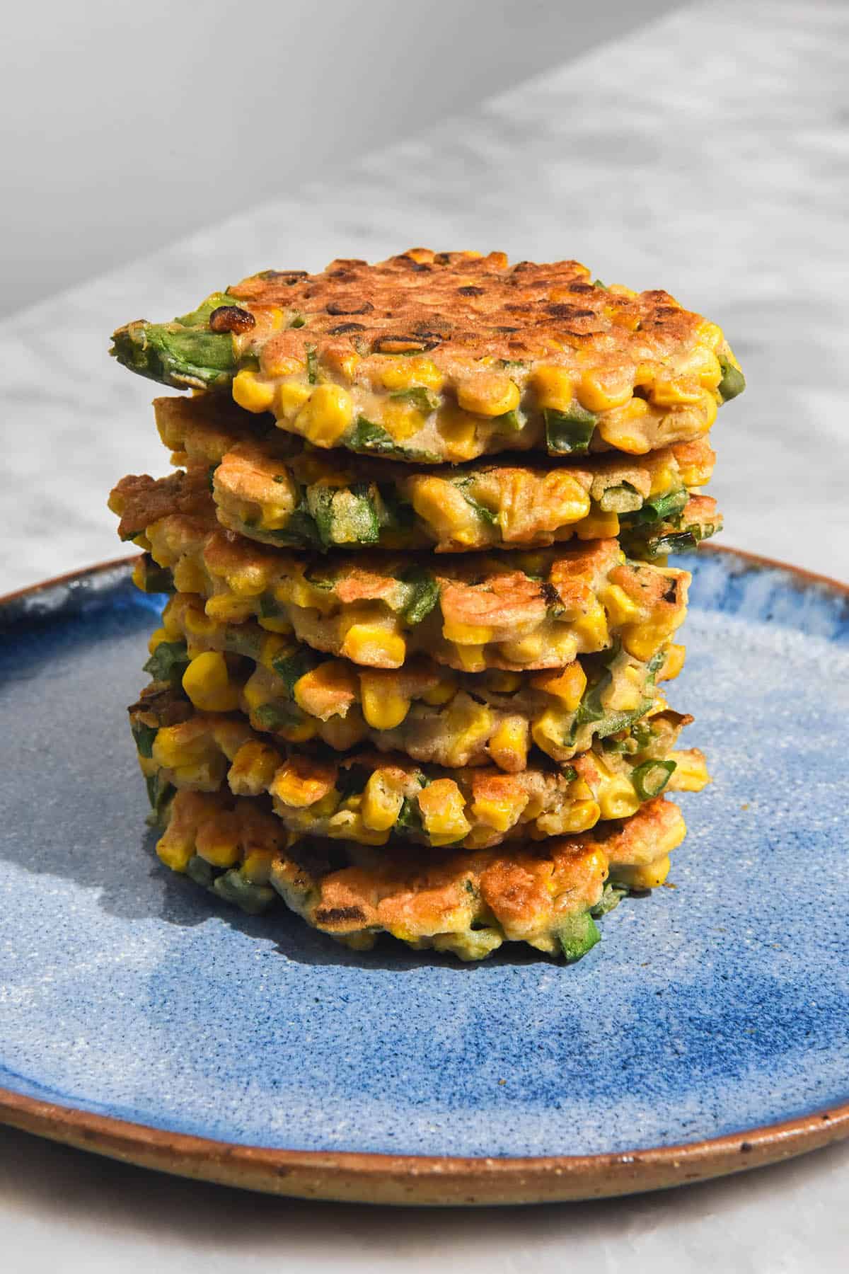 A brightly lit side on image of a stack of gluten free corn fritters on a bright blue ceramic plate atop a white marble table. 