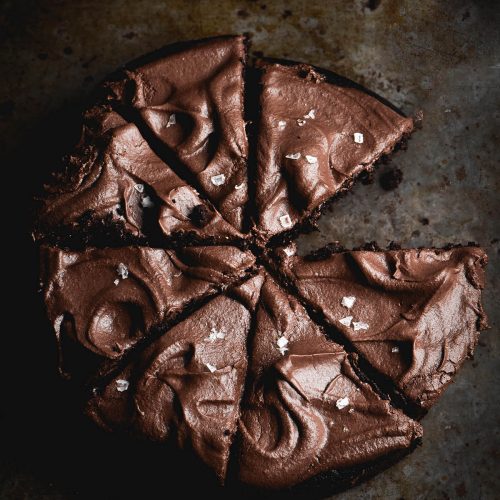 A dark and moody image of a gluten free vegan chocolate cake topped with buttercream and sea salt flakes. The cake has been sliced and one slice has been removed.