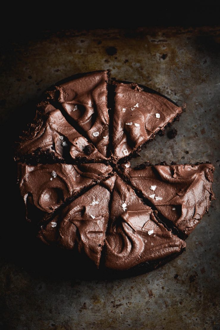A dark and moody image of a gluten free vegan chocolate cake topped with buttercream and sea salt flakes. The cake has been sliced and one slice has been removed.
