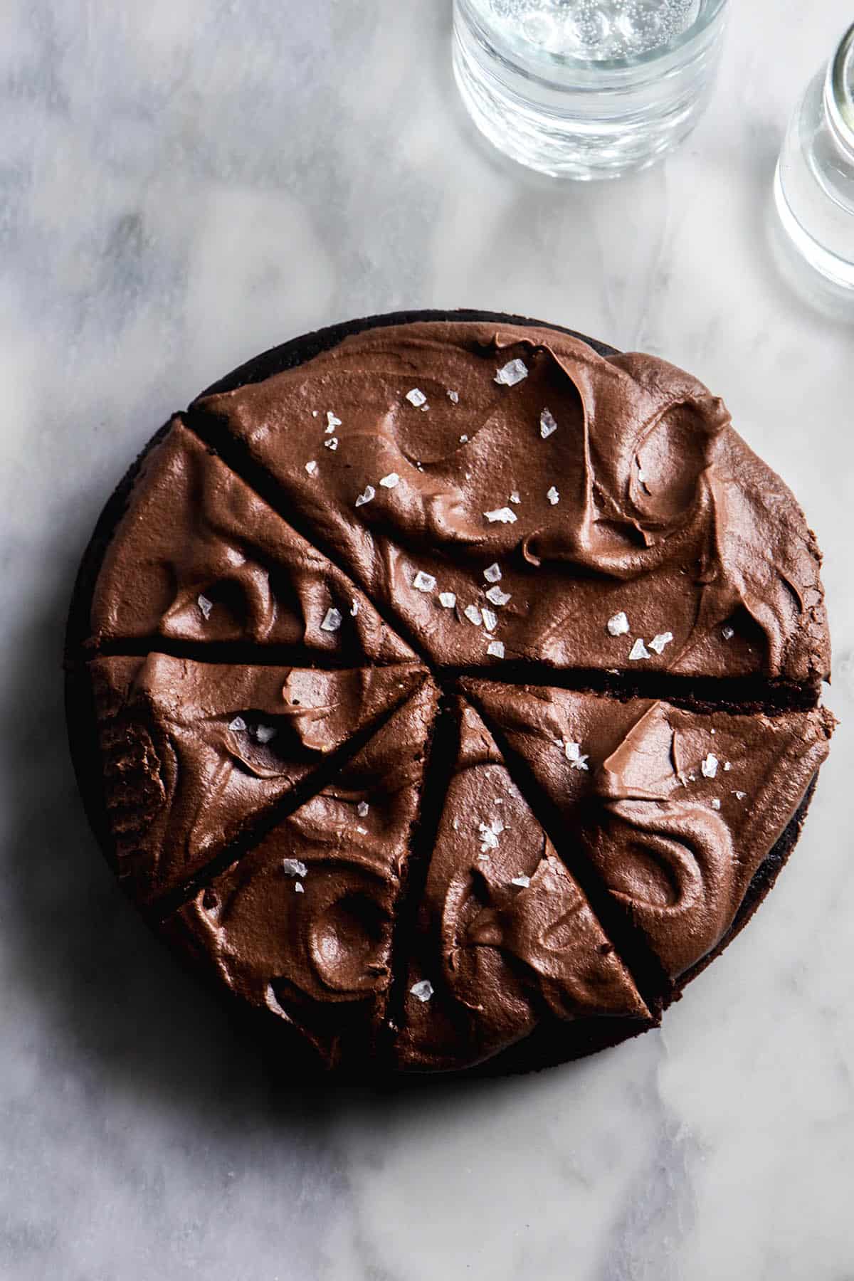 An aerial image of a gluten free vegan chocolate cake on a white marble table. The cake has been topped with vegan chocolate buttercream and a sprinkle of sea salt flakes and is cut into slices.