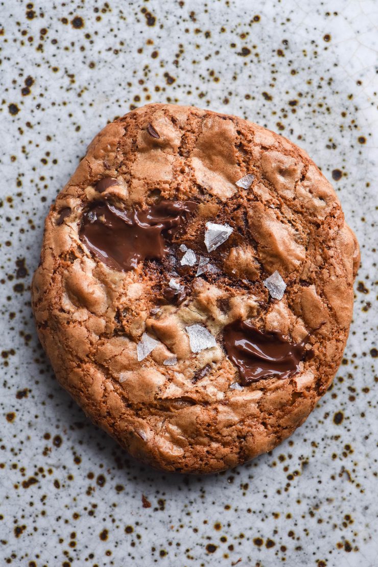 An aerial close up of a grain free tahini choc chip cookie on a white speckled ceramic plate. The cookie has melty chocolate chips beneath a crackly golden surface and is topped with sea salt flakes